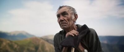 An elderly man stands outside behind mountains