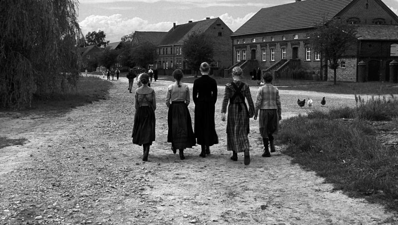 A black and white photo shows a group of children in 1900s dresses walking down a dirt path.