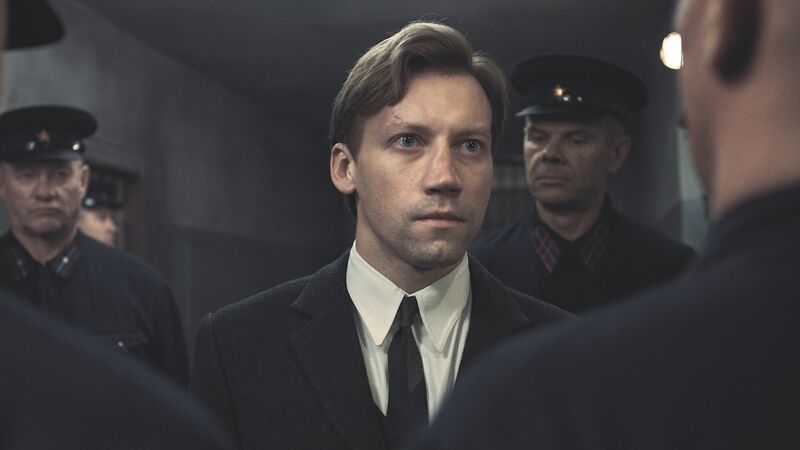 A man in a suit looks at a group of army officers with a determined look on his face