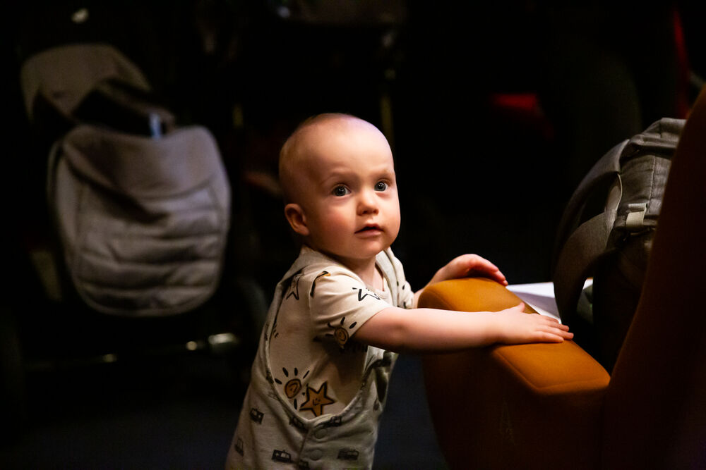 A cute baby standing up, holding on to an orange cinema seat for support.