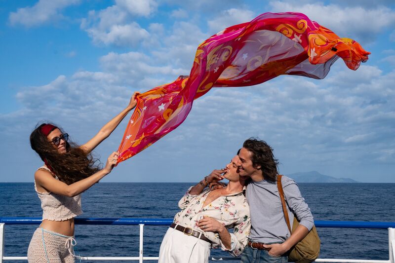 A young woman wearing sunglasses holds a scarf in the air next to the sea, while two men look and smile.