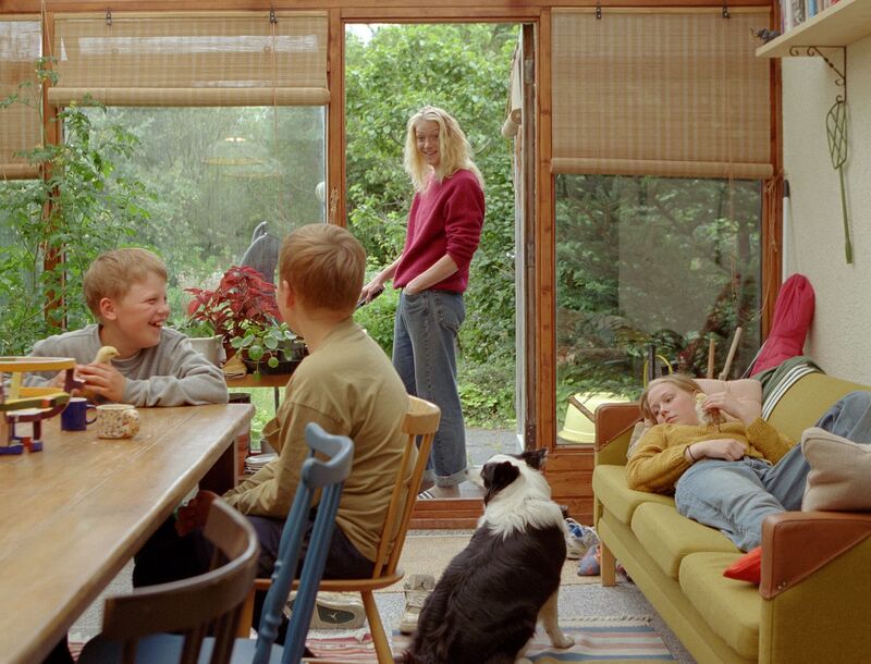 Three children sit in a bright, wooden living room with a green garden in the background. A woman turns round, smiling at them