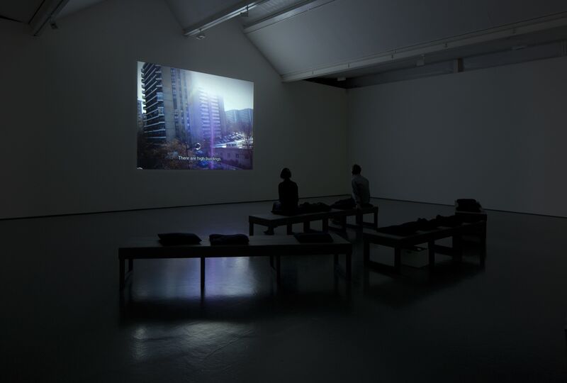At DCA Galleries for Nikolaj Bendix Skyum Larsen's exhibition, two people sit on a bench watching a projection. On the projection, there is a video of a block of flats.