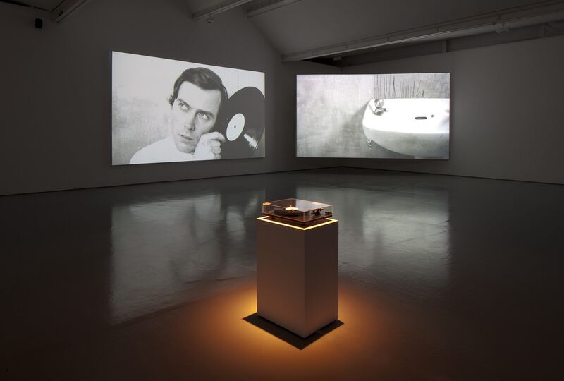From Hiraki Sawa's exhibition. A record player with a glass case its on a plinth in the middle of DCA Galleries. Two projection screens show image - one of a man holding a record up to his ear, the other of a bathroom sink.