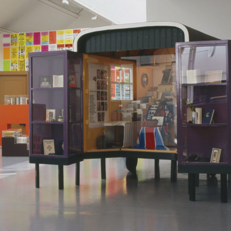 From Allen Ruppersberg's exhibition at DCA. A caravan is blocked off with glass to make a display. Inside, there are cabinets with books, a flag, a potted plant and a poster.