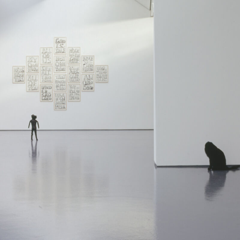From David Shrigley's exhibition in DCA Galleries. A statue of a headless cat can be seen in the foreground. In the background, there is a small black statue of a man. On the walls, there are text-based paintings, arranged in white frames in a diamond shape.