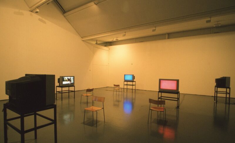 A white gallery space is filled with televisions with blue and pink screens on them, and wooden chairs.