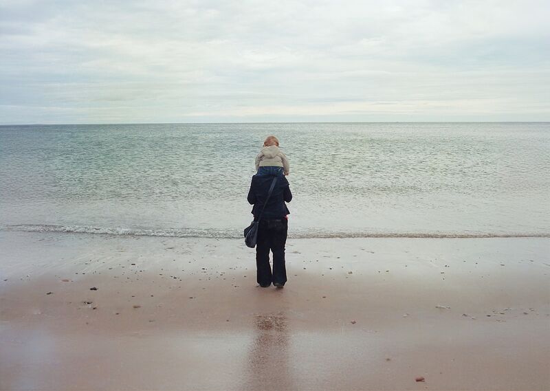 A woman stands on a beach, her young son on her shoulders.