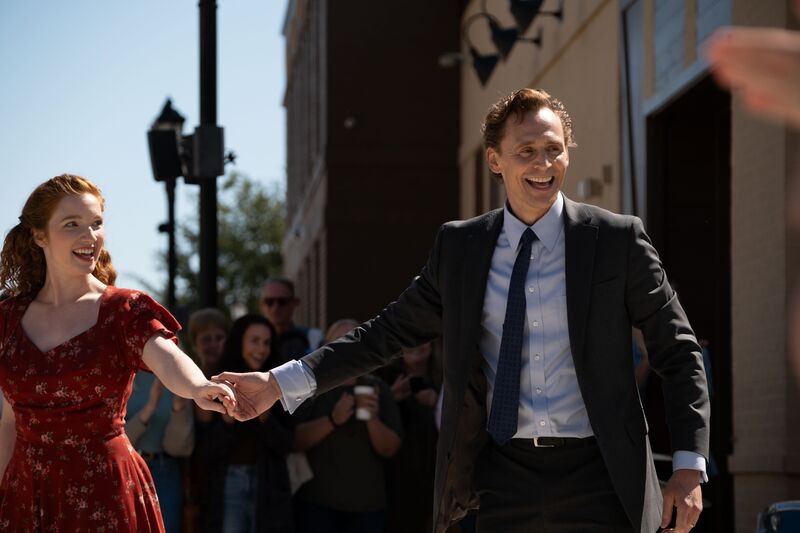 A man wearing a suit and a woman in a red dress hold hands and smile on a city street.