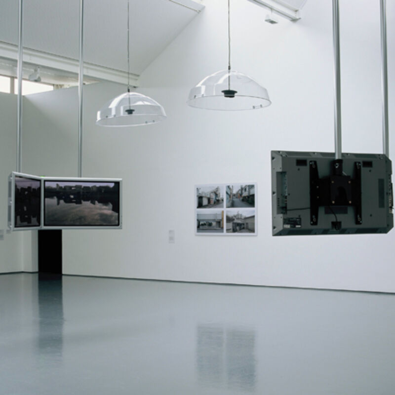 From Behind Closed Doors at DCA Galleries. Glass lights hang from the ceiling, along with flat screen televisions which have black-and-white images on them. Black-and-white photographs can also be seen on the walls.