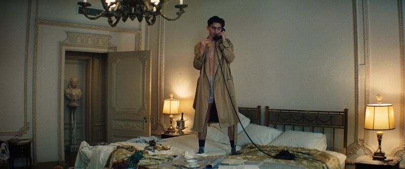A shirtless man in a coat stands on a bed in a luxury hotel room, talking on the phone