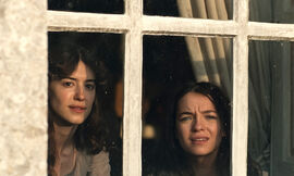 Two dark haired young women look out from a window