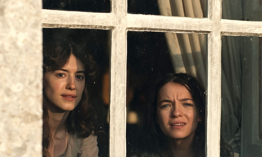 Two dark haired young women look out from a window
