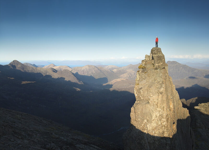 A man stands on top of a very steep rock with a view of an impressive mountain range.