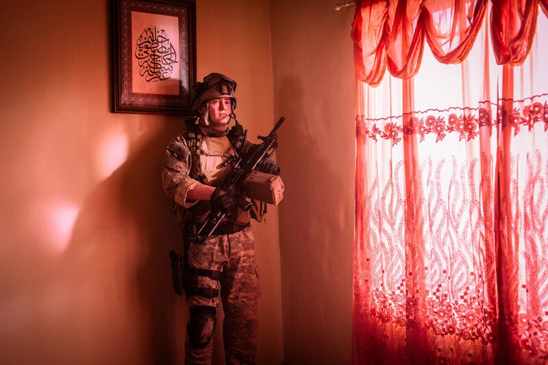A soldier in full camouflage and protective clothes stands in a room with a red curtain.