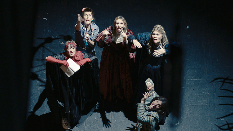 A circle of stage actors gesture wildly up at the camera above them