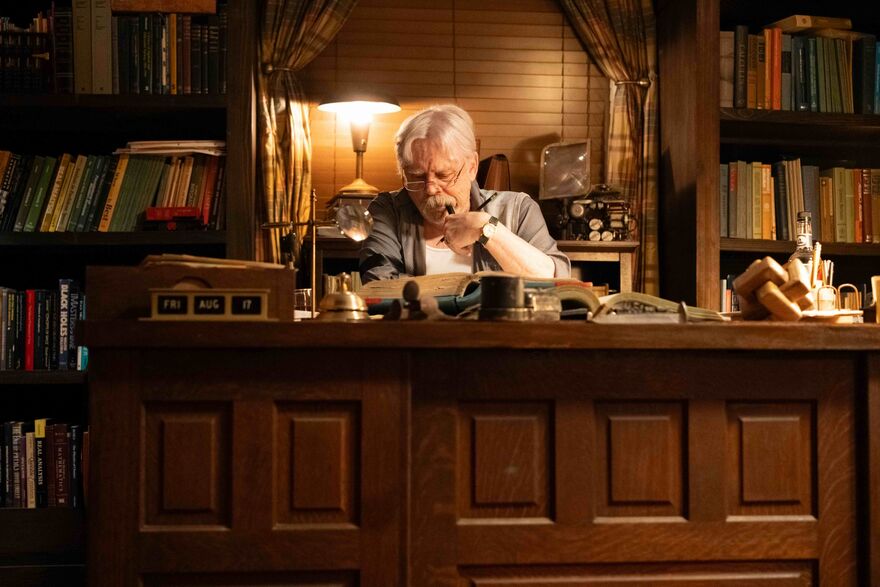 An older man sits at a wooden desk with many books behind him.