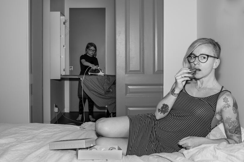 Black and white image of women in glasses eating while lying on a bed and a child in the background does the ironing