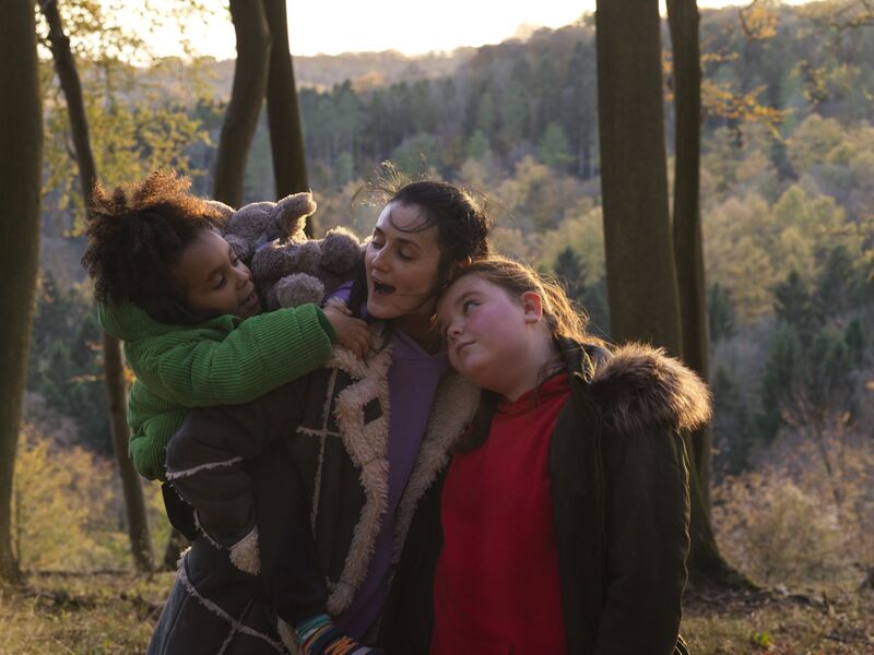 Standing in a forest, a woman hugs two little girls.