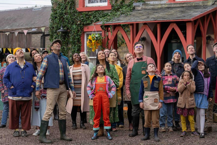 A group of people standing in a village wearing colourful clothes look up.