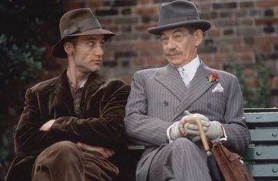 Two men, one in a brown suit and hat, and another in a pinstripe suit and hat, sit together on a park bench.