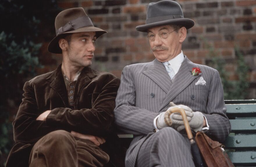 Two men, one in a brown suit and hat, and another in a pinstripe suit and hat, sit together on a park bench.