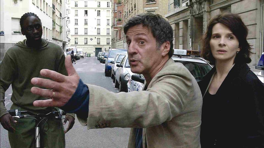 A man stands in the street, looking angry and gesturing with his arm. Two people stand next to each other.
