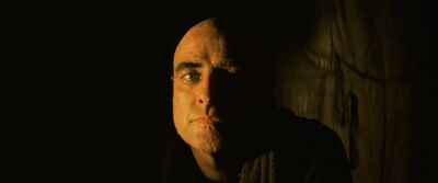 A man with a neutral expression sits, his face is lit from one side.