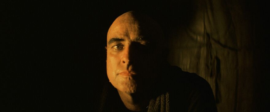A man with a neutral expression sits, his face is lit from one side.