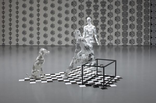 Three white mannequins from Johanna Basford's exhibition. One mannequin of a human is standing, another is sitting. There is also a white mannequin of a dog. All of the figures are decorated with intricate patterns of black ink.