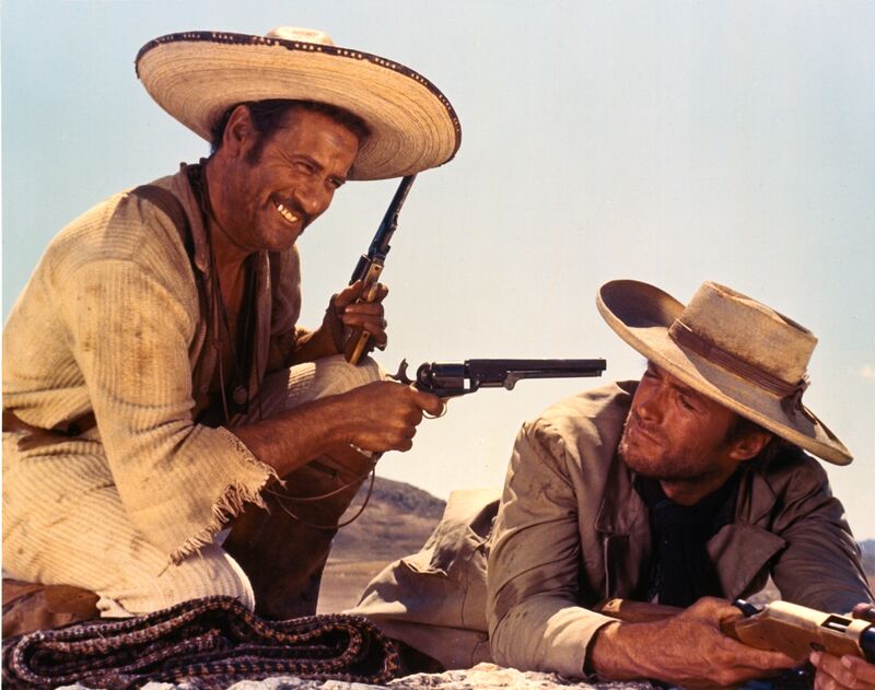 A grinning man holds a gun to the head of a man lying down