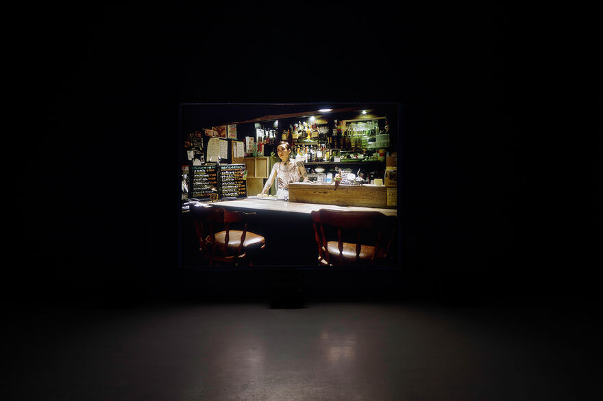 This image shows a dark gallery with a video projection. In the film we can see a young person stood behind a bar, with two empty chairs pulled up on the other side. 