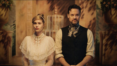 Blonde haired woman in a white dress stands beside a man with short dark hair and a beard. The both stand in front of a backdrop