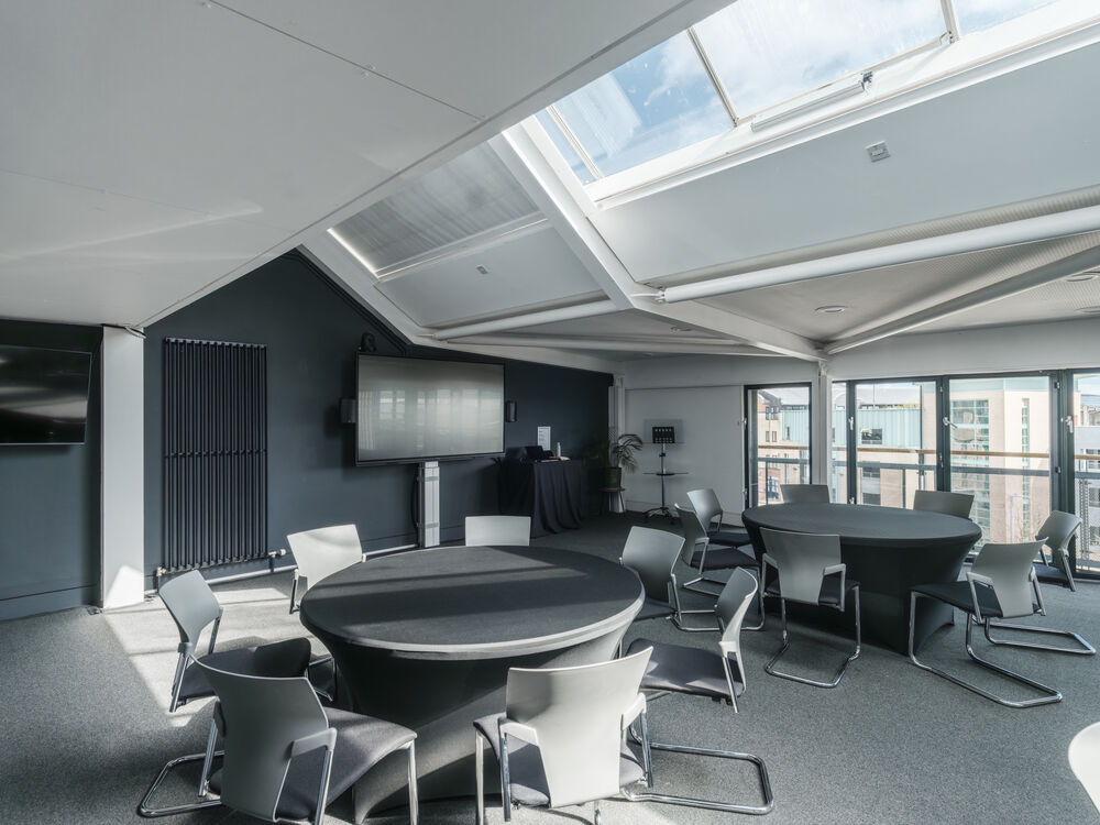 A room with round tables and chairs around them - there are windows in the ceiling and large windows on the far wall. A screen is visible in the background