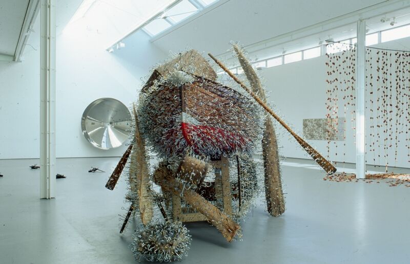 In DCA Gallery, a white fishing boat is covered in metal hooks and nails. In the background, a mobile covered in flowers can be seen, and a large circular mirror.