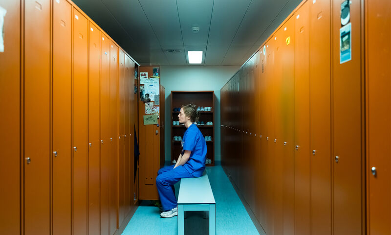 A nurse in a blue uniform sits on a bench in a locker room alone.