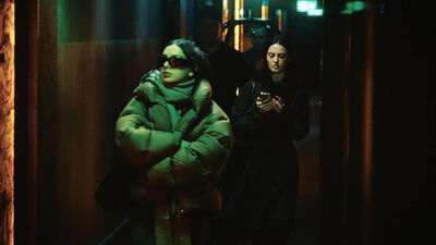 A woman wearing dark sunglasses and a puffer jacket walks down a street in the dark, followed by a woman on the phone