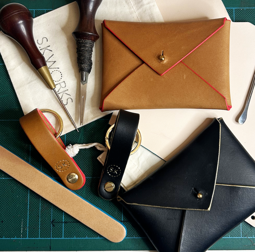 Table with two leather card purses and two keyrings. One of each is black, the other is brown. They are surrounded by leather working tools