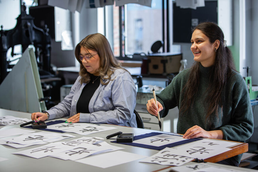 Two people sitting at a desik holding brushes and working on Japanese Calligraphy symbols