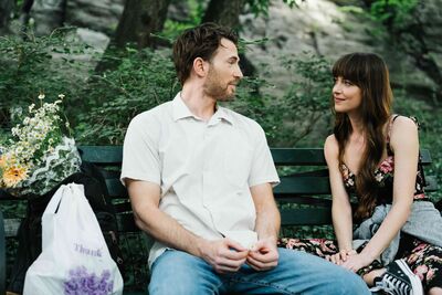 A man and woman sit on a park bench together, next to flowers in a plastic bag, smiling.