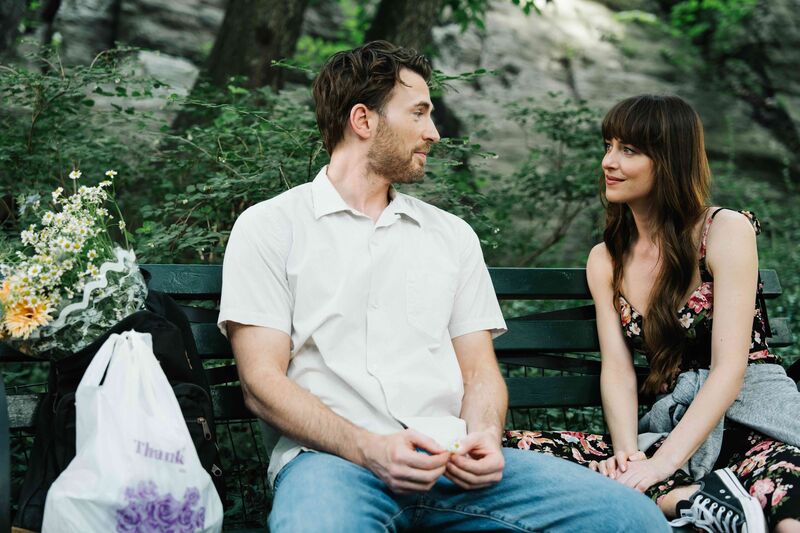A man and woman sit on a park bench together, next to flowers in a plastic bag, smiling.