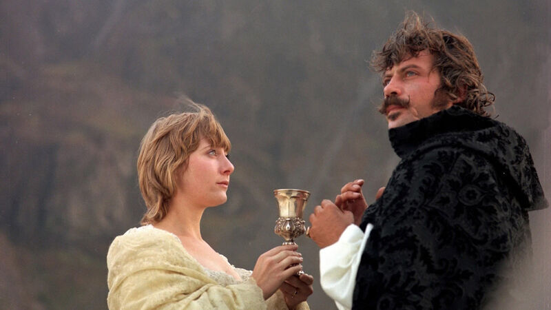 A woman holding out a chalice to a man in a black cape