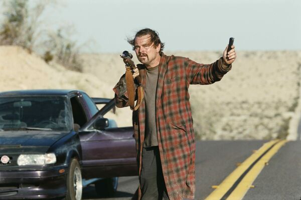 A man in a long tartan dressing gown stands in the middle of a road holding a gun.