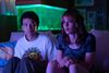Two young people sit next to each other on a sofa with a fish tank behind them. The room is dark, illuminated by the TV. 