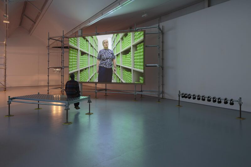 A person sits in DCA gallery on a scaffold platform watching a video. The person on the screen stands in front of a shelving unit with many green boxes on it.