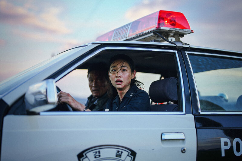 Two police officers sit in a police car, looking out of the window in fear