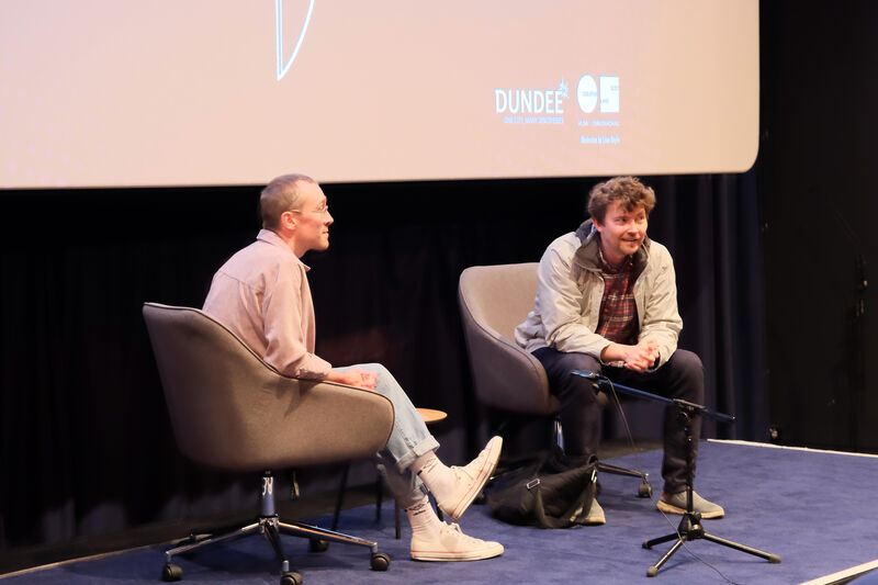 Two people sitting on chairs on a small stage in front of a big screen