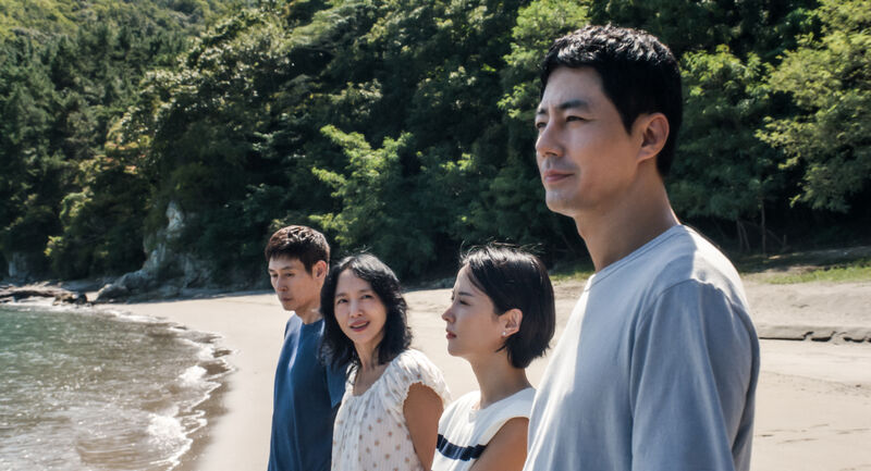 Two couples stand on a beach. One looks out to see, while the woman in the second couple looks over at the other man