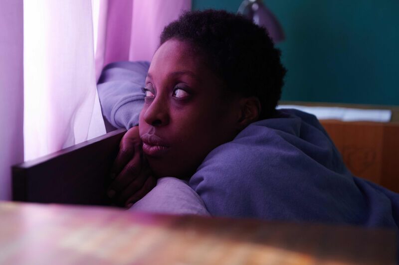 A woman leans on a bed's headboard, looking out a crack in the curtain