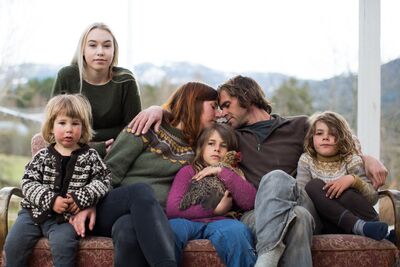 A family with three young children and a teenager sit outside on a couch. The couple have their foreheads together.